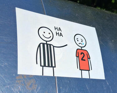 Line drawing of man in black and white striped shirt pointing at a man wearing a red jersey with the number 2 on it. The black and white shirt guy is saying “HA HA” and the man with the red jersey is sad and has a tear coming from his eye.
