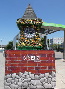 Model of uhrturm covered in flowers. it is standing on a pedestal made to look like a brick wall.