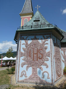 Shield on tree with letters IHS. At bottom left and right are capital alpha and omega. Church spire is in background
