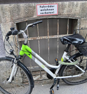 White bicycle with green trim parked in front of a sign reading Fahrräder anlehnen verboten!