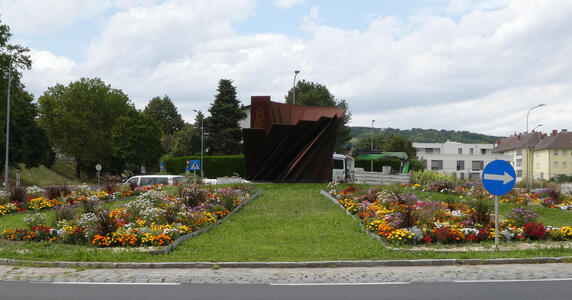 Cylindrical bronze sculpture in middle of traffic circle. Flower bed to either side of the sculpture.