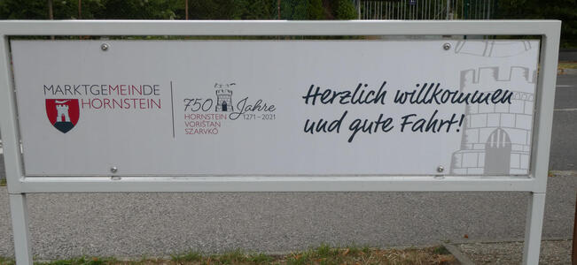 Sign with shield at left. text: Marktgemeinde Hornstein 750 Jahre 1271-2021 Herzlich willkommen und gute Fahrt! In the first part of the text, letters in red spell out MEIN HORNSTEIN