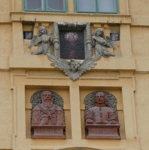 Above castle entrance, two busts of old men. Above them, a frame with a cracked painting, held by angels.