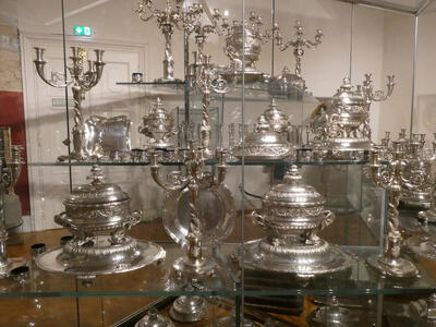 Display case filled with silver teapots serving dishes, and candelabras