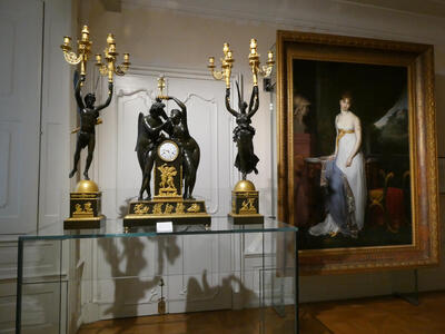 At left, a table with two lamps supported by statues of a man and a woman rspectively. Between them, a desk clock with a man and woman embracing on either side of the clock. At right, a painting of a woman in a white dress with a gold belt