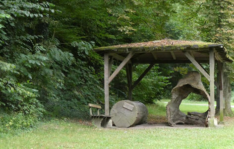 A wooden shelter; under it, a log and a portion of a tree. At left, a small bench