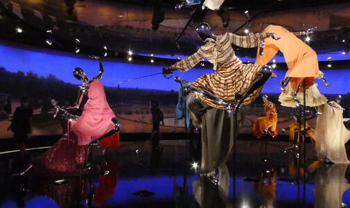 Manikins in dresses as if floatig in air; one is holding an epee.
