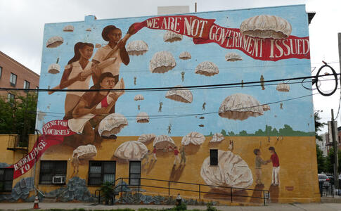 Young women, one holding diploma, another a paintbrush with a strip of paint reading “We are not government issue”. Multiple parachutes in background with text such as Our humanity, our futures, our dreams