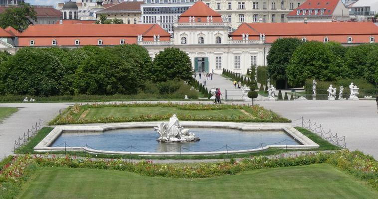 In foreground, a pool with statue in middle. In background, a large white building with red roof.