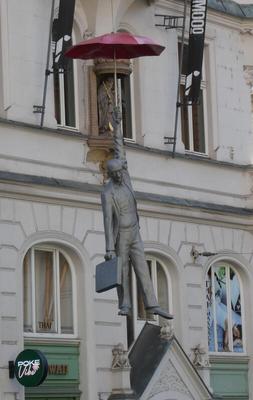 Statue of man with briefcase in one hand. He is hanging from a red umbrella in the other hand.