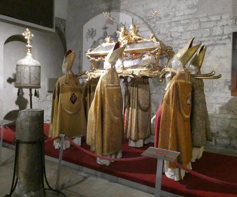 Six mannequins in bishop robes and mitres bearing a coffin with gold statuary atop it.