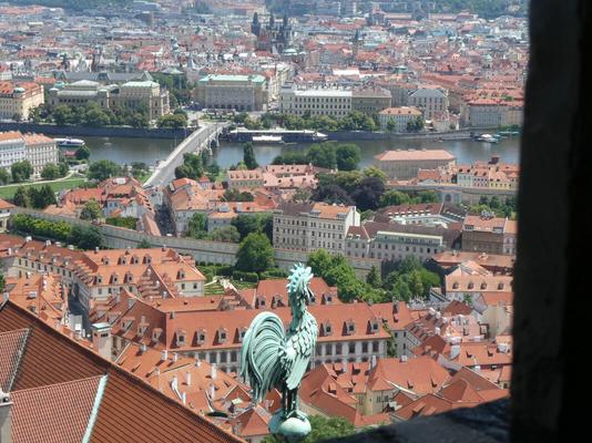 View of city; a bronze rooster attached to the church is in foreground. River and bridge are in midground.