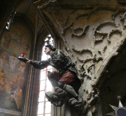 A column with a wood carving of a man in red pants and gray shirt. he is holding a tray with a candle holder on it.