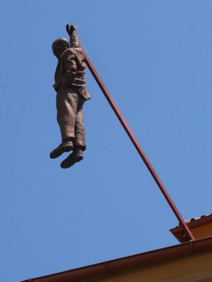 Sculpture of man hanging by one hand from a rod projecting from a ceiling