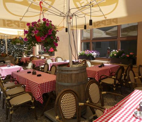 Outdoor café with basket of read flowers hanging from a patio umbrella. Tables are covered with red checkered tablecloths.