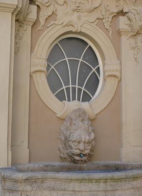 Fountain with spout shaped like a half-human half-fish head. Above it, an oval window with arcs to segment the glass